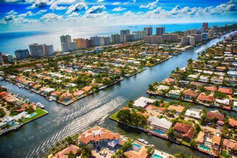 Vibrant aerial view of Pompano Beach, Florida, showcasing a network of residential canals lined with waterfront homes, boat docks, and lush greenery, with a backdrop of high-rise oceanfront buildings and the Atlantic Ocean under a bright blue sky.