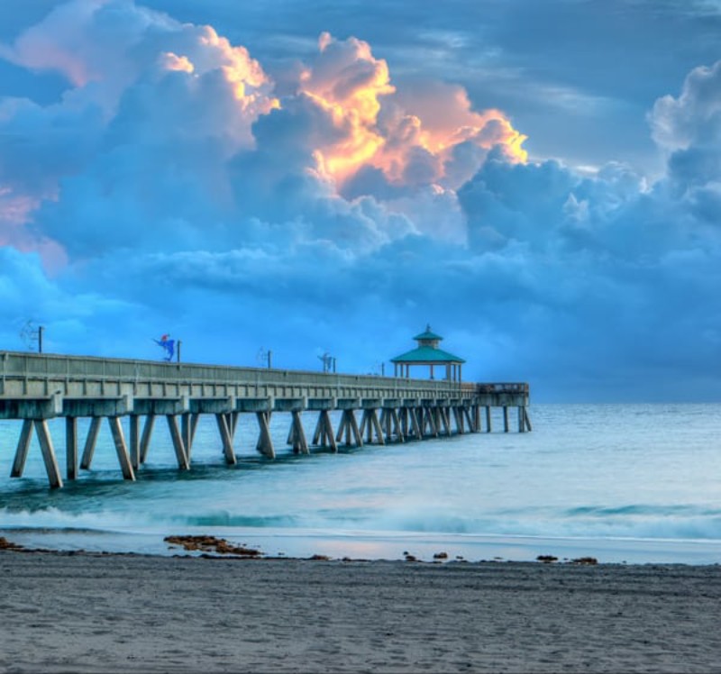 Serene view of Deerfield Beach pier stretching into the ocean during sunrise or sunset, with a dramatic sky of glowing orange and blue clouds above calm waves and a sandy beach in the foreground.