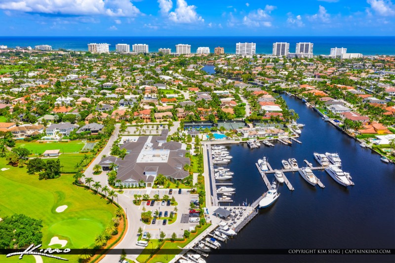 Aerial view of Royal Palm Yacht and Country Club in Boca Raton, Florida, showcasing a luxury marina filled with yachts, a large clubhouse, manicured golf course, and upscale homes, with the Atlantic Ocean and high-rise buildings in the distance.
