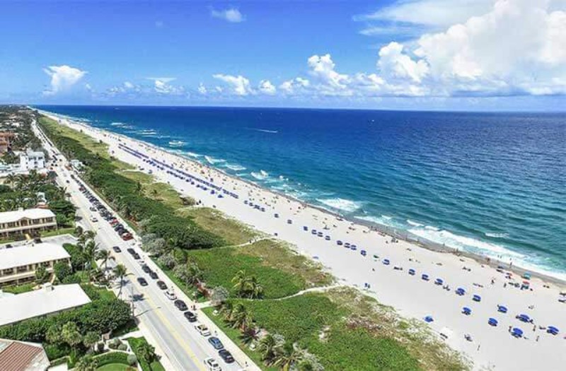 Aerial view of Delray Beach, Florida, featuring a wide sandy shoreline lined with rows of blue umbrellas and beachgoers, with a scenic coastal road and low-rise buildings on the left, all set against a backdrop of turquoise ocean and partly cloudy skies.