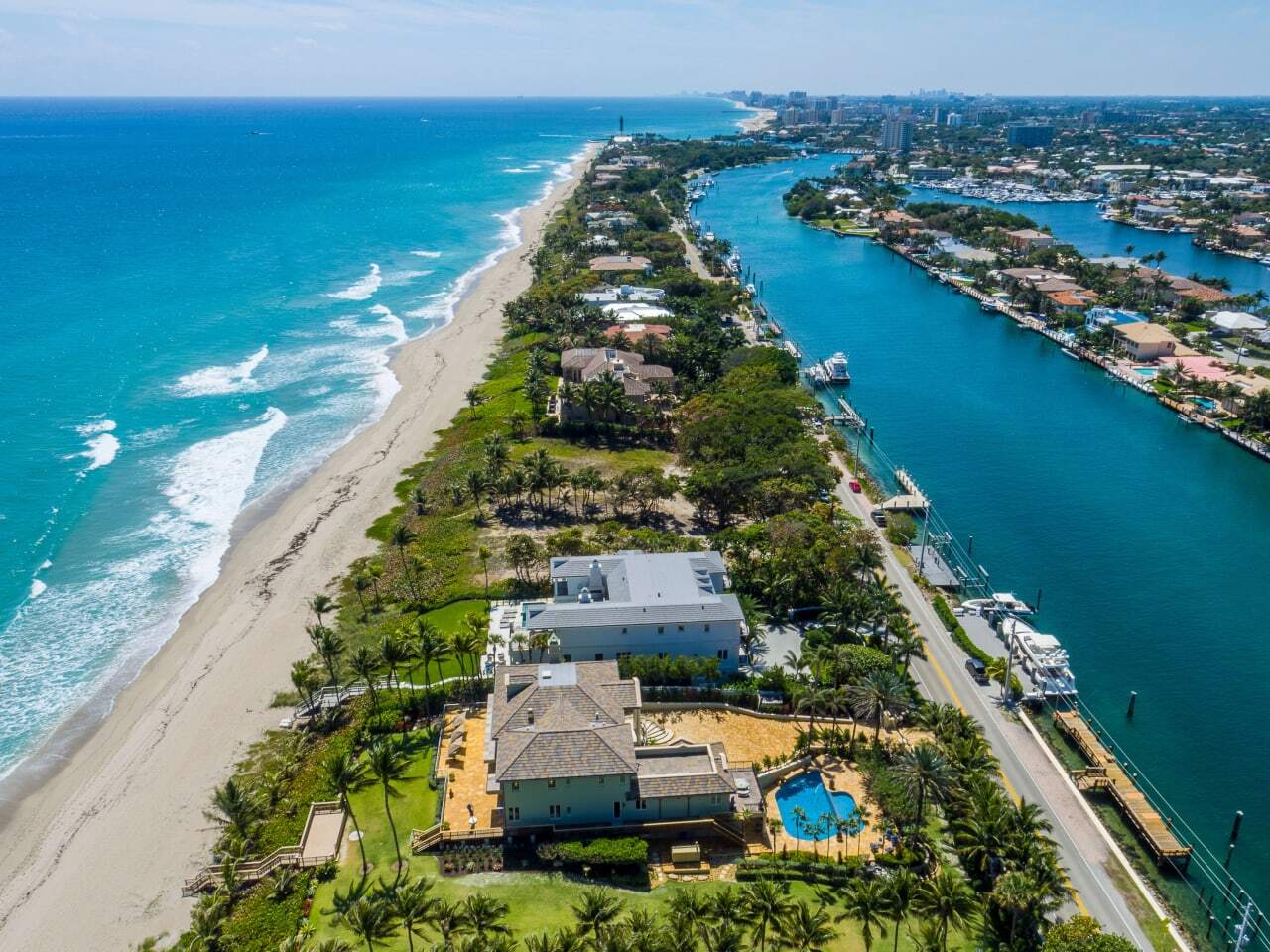 Aerial view of Hillsboro Mile showing a narrow strip of luxury homes between the Atlantic Ocean and Intracoastal Waterway, with turquoise water on both sides, lush landscaping, and a road running down the center.